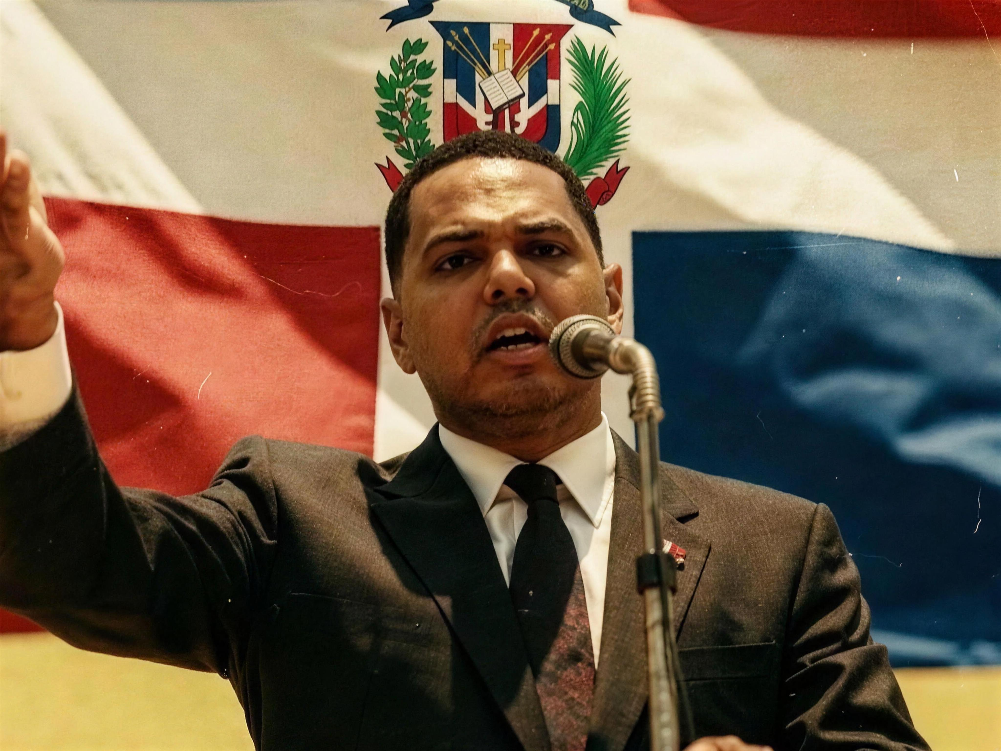 Santiago Matías hablando frente a un micrófono con la bandera dominicana al fondo